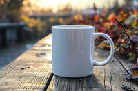 White ceramic coffee mug on rustic wooden table outdoors with autumn leaves and golden sunlight in background, creating cozy morning atmosphere.