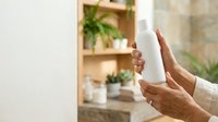 Mature woman with medium skin tone gesturing stop or pause with both hands, wearing neutral beige clothing, indoor setting with wooden shelving and potted plants in background.