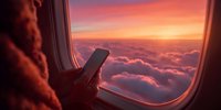 Person using smartphone by airplane window with stunning sunset view over clouds, creating warm orange glow across sky during flight.