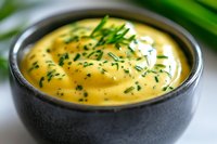Creamy hollandaise sauce garnished with fresh chopped chives in dark ceramic bowl, close-up view showing smooth texture and rich yellow color.