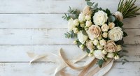 Elegant bridal bouquet with cream and blush roses, eucalyptus leaves and silk ribbon on rustic white wooden background for wedding celebrations.