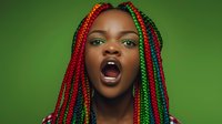 Young African woman with vibrant red and green braids expressing intense emotion against green background, mouth wide open in passionate expression.