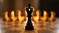 Black chess king piece standing prominently on wooden board with blurred golden chess pieces in background creating dramatic lighting and strategic game atmosphere.