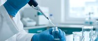 Scientist in blue gloves and lab coat pipetting clear liquid into glass vial in modern laboratory setting with blurred equipment background.