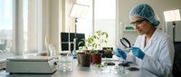 Female scientist in lab coat and surgical cap examining plant seedlings with magnifying glass in bright modern laboratory for botanical research studies.