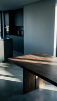 Solid wood dining table with natural grain in a modern dark interior, sunlight casting shadows on concrete floor and matte walls.