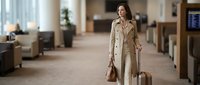 Professional businesswoman in beige trench coat walking through modern airport terminal with luggage, ready for business travel and corporate trips.