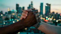 Two diverse hands clasping in a strong handshake against a glowing city skyline at dusk, symbolizing unity, partnership, and multicultural solidarity.