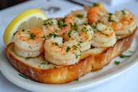 Garlic butter shrimp served on toasted baguette slice with fresh herbs and lemon wedge garnish, close up view of appetizing seafood bruschetta on white plate.