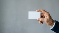 Mature male hand holding blank white business card against neutral gray background for corporate branding and professional networking purposes.