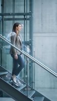 Professional businesswoman in gray blazer walking up modern glass staircase in corporate office building. Contemporary workplace architecture with transparency.