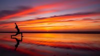 Young woman practicing yoga warrior pose on wet beach sand during dramatic sunset with vibrant orange and purple sky colors reflecting in water.