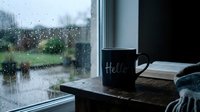 Dark mug with Hello text on wooden table by rain-covered window, open book and cozy scarf beside it, moody autumn atmosphere.
