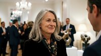 Mature businesswoman with gray hair smiling during corporate networking event in elegant venue with colleagues in background for professional meetings.