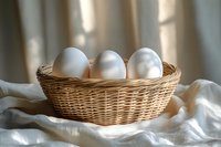 Fresh white chicken eggs in woven wicker basket on soft white fabric with natural window light and shadows creating cozy rustic atmosphere.