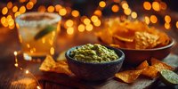 Fresh guacamole dip in rustic bowl with tortilla chips and margarita cocktail on wooden table against warm bokeh lights for festive party atmosphere.