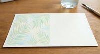 Watercolor tropical palm leaves pattern on white paper lying on wooden desk with glass of water and pencil for creative design projects.