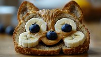 Playful breakfast toast shaped as cat face with banana slices for eyes, blueberries and peanut butter creating cute animal food art for children's meal.