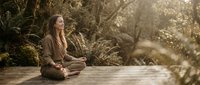 Young woman practicing mindfulness meditation outdoors on wooden deck surrounded by lush tropical plants in warm golden sunlight for wellness and relaxation.