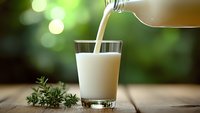 Fresh milk pouring into glass on wooden table with green herbs, healthy dairy beverage for breakfast and nutrition concepts.