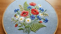 Embroidered wildflower bouquet with poppies, daisies and cornflowers in wooden hoop on light blue fabric. Handmade needlework craft for home decor projects.