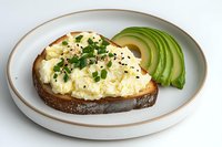 Creamy scrambled eggs on dark toasted bread garnished with fresh herbs and black sesame seeds, served with sliced avocado on white ceramic plate with gold rim.