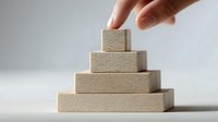 Hand stacking wooden blocks in pyramid shape against neutral background, demonstrating hierarchy, growth, or business strategy concept.