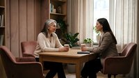 Professional counseling session with mature Caucasian therapist and young Asian woman client discussing mental health in warm modern office setting.
