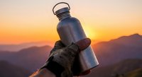 Hiker holds stainless steel water bottle against mountain sunset backdrop. Adventure gear for outdoor activities, camping, and hiking expeditions.