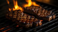 Juicy grilled steaks with perfect grill marks cooking over open flames on barbecue grate, close-up view showing seared meat texture and fire flares.