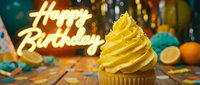 Festive birthday cupcake with yellow frosting swirl, neon Happy Birthday sign glowing in background, colorful confetti and party decorations on wooden table.