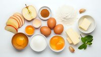 Fresh baking ingredients arranged on white background including eggs, flour, butter, milk, honey and sliced apple for homemade cooking and pastry preparation.
