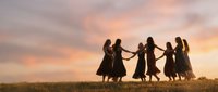 Group of women holding hands in circle dancing together at sunset in field with golden sky creating silhouettes for friendship and unity concepts.
