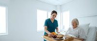 Young female nurse in teal scrubs serving healthy meal to elderly Caucasian woman patient in bright hospital room for healthcare nutrition support.