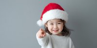 Smiling Asian girl wearing red Santa hat pointing finger at camera against gray background, festive holiday portrait in white sweater.