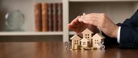Businessman hands protecting wooden house models on desk with books in background symbolizing real estate insurance and property protection services.