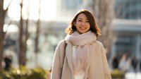 Young Asian woman in beige coat and pink scarf smiling outdoors in urban setting during golden hour light for lifestyle and fashion content.