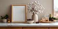 Elegant winter home decor with cotton branches in ceramic vase, gold frame, pine cones, and miniature wooden house on marble shelf against beige wall.