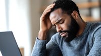 Stressed African American man with beard holding head in hand while working on laptop, experiencing work pressure and mental fatigue in modern office environment.