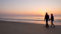 Couple walking on beach at sunset with golden sky and ocean waves creating romantic silhouette scene for travel and relationship concepts.