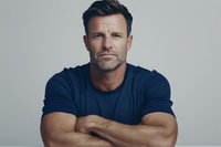 Confident mature Caucasian male with crossed arms wearing navy t-shirt against light background, displaying serious expression and athletic build. Professional headshot style.