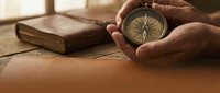 Hands holding vintage brass compass with old leather-bound book on wooden table in warm sunlight for navigation and adventure concepts.
