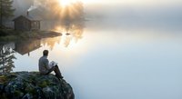 Solitary man sitting on rocky shore contemplating misty lake at sunrise with rustic cabin and boat creating peaceful morning atmosphere for mindfulness content.