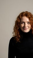 Young Caucasian woman with curly red hair smiling warmly in black turtleneck sweater against neutral beige background for professional headshot photography.