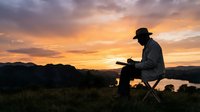 Silhouetted male farmer in work clothes and hat sitting on equipment at sunset, reviewing crop data on laptop in rural agricultural field with golden sky and mountains.