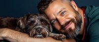 Smiling bearded mature man in green sweater cuddling with adorable mixed breed dark-haired dog in close-up portrait showing bond between pet and owner.