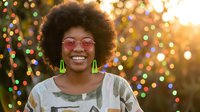 Happy young African American woman with afro hairstyle and pink sunglasses smiling at Christmas lights background during golden hour sunset.