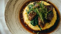 Braised beef short rib served on creamy polenta with sautéed spinach, rich gravy, and fresh rosemary garnish on rustic ceramic plate.
