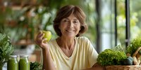 Smiling mature Caucasian woman holding green apple in bright kitchen surrounded by fresh vegetables promoting healthy eating and wellness lifestyle.