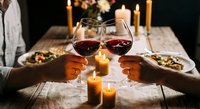 Romantic dinner date with couple toasting red wine glasses by candlelight on rustic wooden table with gourmet meal for anniversary celebration.